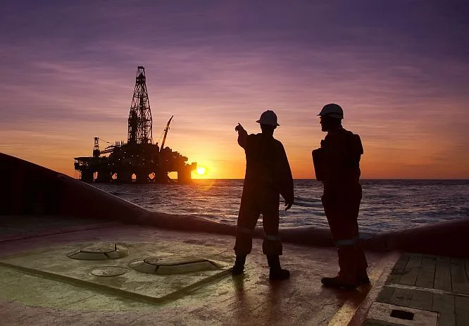 Two people in PPE on a vessel deck looking toward an offshore rig at sunset